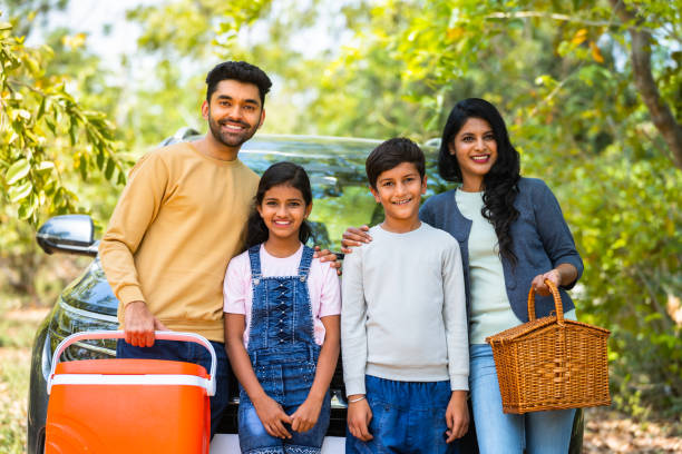 Happy smiling family with sibling kids standing in front of car for picnic by looking camera during holiday travel - concept of joyful lifestyle, family time and freedom.